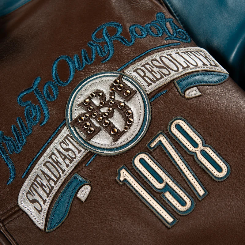 Close-up of the Pelle Pelle True To Our Roots brown jacket showing teal embroidery, decorative crest, cream lettering, and metallic-tone studs