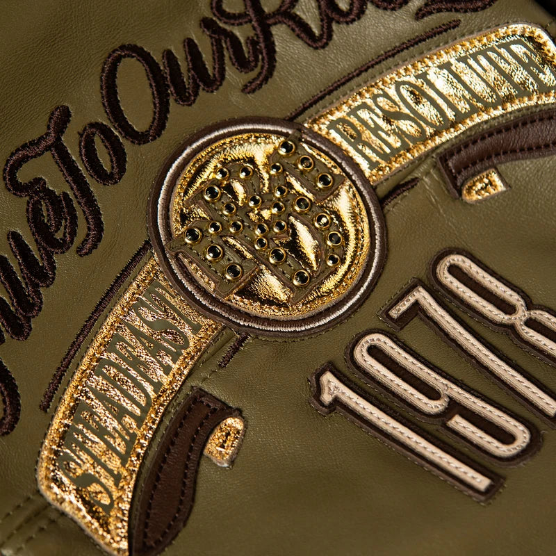 Close-up of the olive green Pelle Pelle True To Our Roots jacket showing gold-tone crest details, decorative studs, and embroidered lettering
