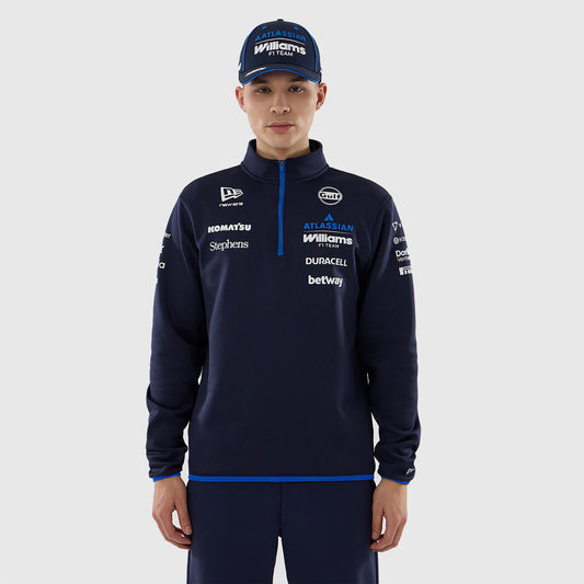 A man wearing a navy blue Williams Racing team quarter-zip pullover and matching cap, featuring sponsor logos such as Homats, Saporta, and Duracell, against a light gray background.
