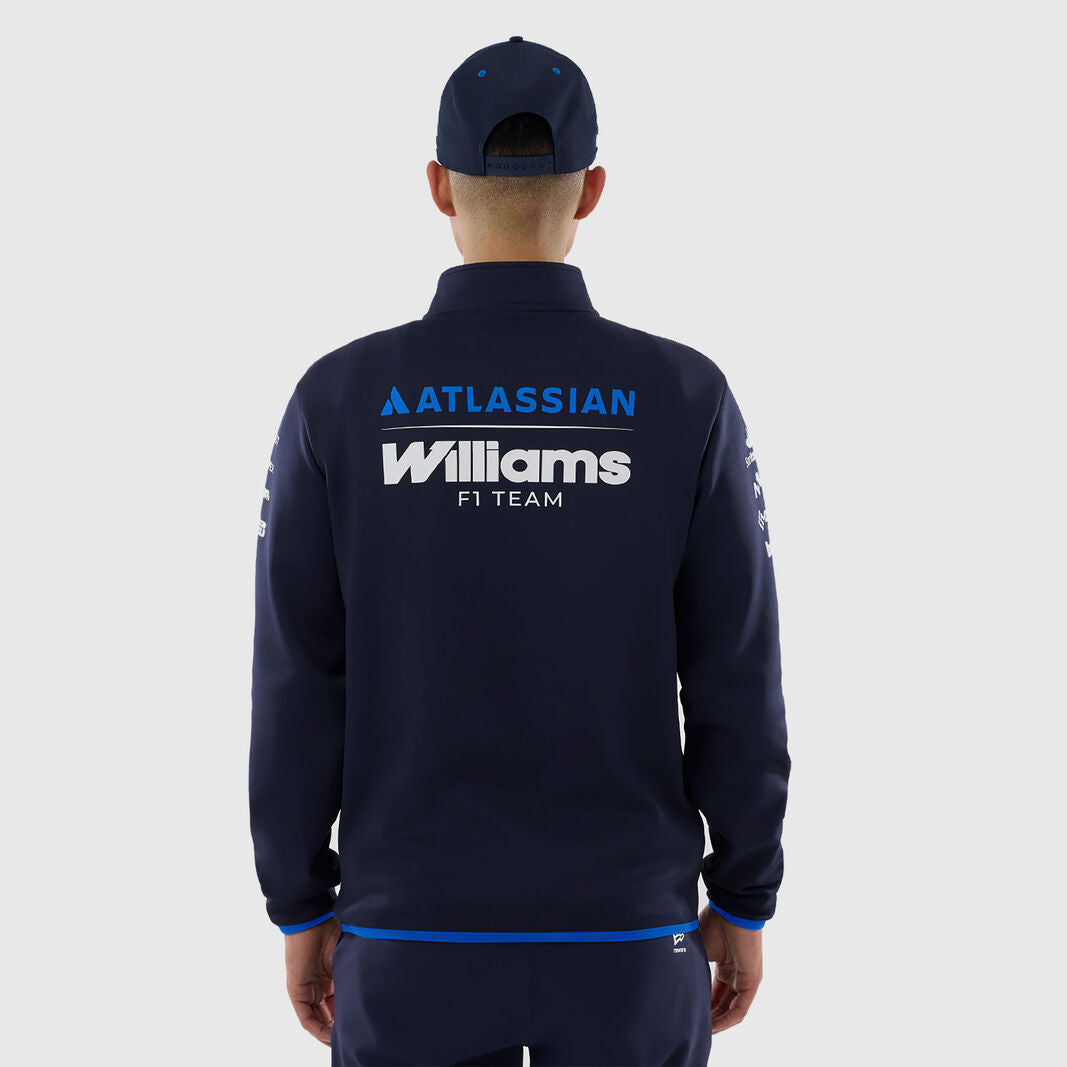 A man viewed from the back wearing a navy blue zip-up jacket with 'ATLASSIAN Williams F1 TEAM' printed in white and blue on the back, along with a matching navy cap, against a light gray studio background.