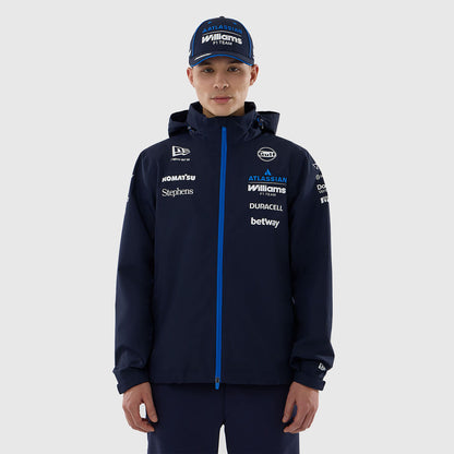 A man wearing a dark navy blue hooded jacket with a bright blue zipper and multiple sponsor logos, paired with matching navy pants and a navy cap, photographed against a light gray studio background.