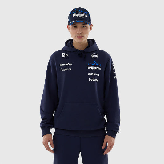 A man wearing a navy blue Williams Racing pullover hoodie and matching navy pants, paired with a navy cap, against a light gray studio background. The hoodie features multiple white and blue sponsor logos including Williams, Castor, and Betway.