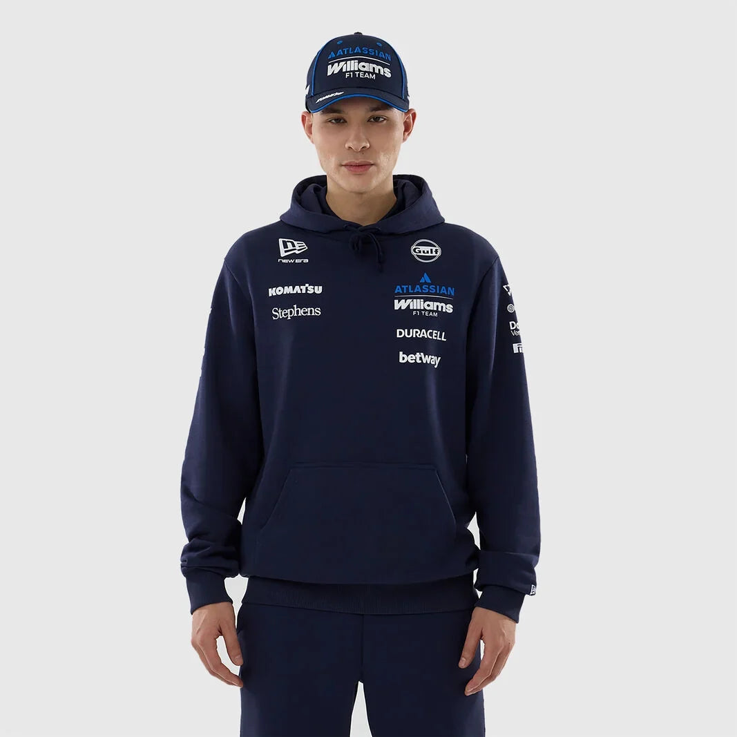 A man wearing a navy blue Williams Racing pullover hoodie and matching navy pants, paired with a navy cap, against a light gray studio background. The hoodie features multiple white and blue sponsor logos including Williams, Castor, and Betway.